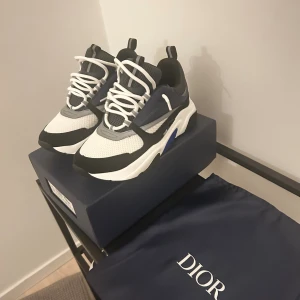 Dior B22 sneakers i svart, vit och blå - Säljer ett par Dior B22 sneakers med chunky sula i svart, vit och marinblå. Skorna har meshpaneler, detaljer i skinn och syntet samt vita snören. Snygg sportig design med tydlig Dior-logga på sidan och insidan. Perfekta för dig som gillar lyxiga statement-sneakers. Sprillans nya som är oanvända. Kontakta för frågor eller bud!