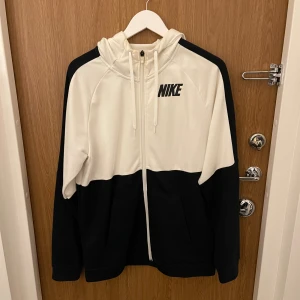 Nike hoodie i svart och vitt - Nike hoodie med dragkedja i svart och vitt. Tröjan har huva med snörning, två fickor framtill och klassisk Nike-logga på bröstet. Perfekt för en sportig och avslappnad stil.