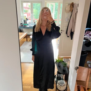 Svart omlottklänning  - Snygg svart långklänning i omlottmodell med v-ringning och knyt i midjan. Klänningen har långa ballongärmar med breda muddar och är gjord i ett glansigt, satinliknande material som ger en lyxig känsla. Normal i storlek! 