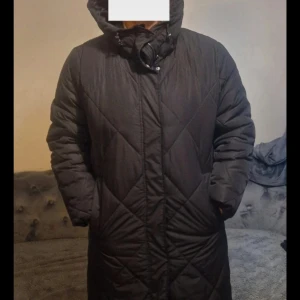 Svart lång pufferjacka Vero Moda S - Lång svart pufferjacka från Vero Moda med quiltad design och huva. Jackan har dragkedja och knappar framtill, samt två sidofickor. Perfekt för kalla dagar och ger en snygg siluett med sin raka passform.