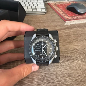 Omega x Swatch Speedmaster MoonSwatch - Omega x Swatch Speedmaster MoonSwatch Mission to the Moon med svart urtavla, vita detaljer och svart textilrem. Klockan har biokeramisk boett, kronograf och tachymeter. En ikonisk modell med cool design och tydliga kontraster.