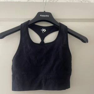 Svart ribbad linnetopp - Snygg svart ribbad linnetopp med sportig design och coola korsade band i ryggen. Toppen är ärmlös och har en tight passform som passar perfekt till träning eller chill. Materialet är mjukt och stretchigt för maximal komfort.