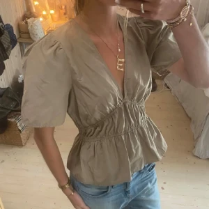 Beige blus med puffärm från Modström - Säljer en beige blus från Modström med v-ringning, puffiga korta ärmar och markerad midja med rynk. Blusen har en lätt volang nertill och är tillverkad i ett tunt bomullstyg. Perfekt att styla med jeans eller kjol. Köpt för 950kr