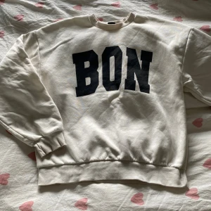 Vit sweatshirt med svart BON-tryck - Säljer en oversized vit sweatshirt med stort svart BON-tryck på bröstet. Tröjan har rund halsringning, ribbade muddar vid ärmslut och nederkant. Perfekt för en chill och avslappnad stil.