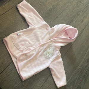 Ljusrosa hoodie med nalle från KappAhl - Supersöt ljusrosa hoodie för baby från KappAhl med broderad nalle på bröstet, knappstängning framtill och en liten ficka. Mjuk och skön i bomullsmix, perfekt för mysiga dagar. Har huva och långa ärmar.