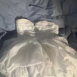 Vit axelbandslös kortklänning - Supersöt vit axelbandslös kortklänning med hjärtformad urringning och fluffig kjol i flera lager. Klänningen har en snygg, figurnära överdel med rynkade detaljer och en voluminös kjol som ger en riktigt drömmig känsla. Perfekt för dig som vill sticka ut och känna dig extra fin!