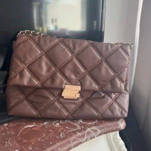 Quiltad brun axelväska med kedja - Snygg brun axelväska med quiltat mönster och guldfärgad kedja. Väskan har ett elegant spänne framtill och är perfekt för dig som gillar stilrena accessoarer. Tillverkad i mjukt material med lyxig känsla.