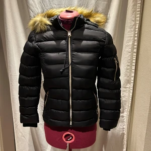 Svart jacka från Vinson Polo Club - Svart pufferjacka från Vinson Polo Club med fuskpäls på huvan. Jackan har guldfärgade dragkedjor, ribbade muddar och en patch på ärmen. En jacka för kalla dagar med sin vadderade design och snygga detaljer.