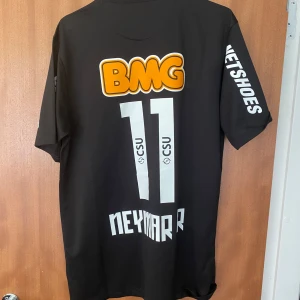 Santos Neymar 11 - Oanvänd Santos FC fotbollströja med Neymar 11 på ryggen. Svart med vita och gula tryck, klubbmärke på bröstet och flera sponsorloggor. Tillverkad i Dri-FIT-material som andas, perfekt för match eller träning. Klassisk passform och korta ärmar.