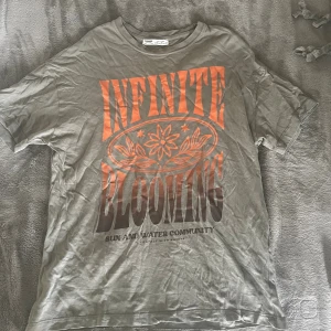 Kaki färgad t-shirt Pull&Bear Infinite Blooming XS - Kaki färgad t-shirt från Pull&Bear i storlek XS med tryck i orange och svart text 'INFINITE BLOOMING' och blommotiv på framsidan. Klassisk rund hals och korta ärmar. Skön och mjuk bomullskvalitet. Använd fåtal gånger. Skriv vid fler frågor!!💓