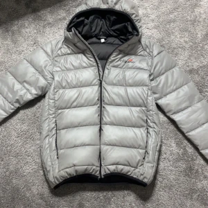 Grå pufferjacka från Ellesse - Säljer en stilren grå pufferjacka från Ellesse med huva och svart innerfoder. Jackan har quiltad design, dragkedja framtill och Ellesse-logga på bröstet. Perfekt för kyliga dagar och enkel att matcha med det mesta. Knappt använd, köpt från zalando för 1400.