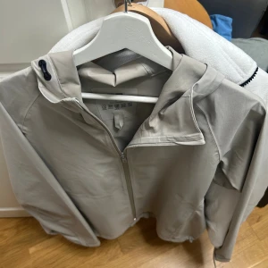 Beige vindjacka från Uniqlo med huva - Säljer en stilren beige vindjacka från Uniqlo i storlek M. Jackan har huva, dragkedja framtill och är tillverkad i ett lätt och stretchigt material. Perfekt för blåsiga dagar och enkel att matcha med olika outfits.