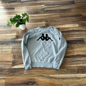 Grå sweatshirt från Kappa - Grå sweatshirt från Kappa med det klassiska svarta logotrycket på ryggen. Tröjan har rund halsringning och långa ärmar. Mjuk insida och avslappnad passform, perfekt för en chill och sportig stil.