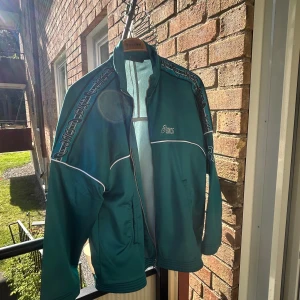 Grön vintage Asics vindjacka tracktop (M) - Retro grön vindjacka från Asics med vita detaljer och logga på bröstet och ärmarna. Jackan har dragkedja framtill, ståkrage och två fickor. Snygga kontrastlinjer och tryck längs axlarna ger en sportig vibe. Perfekt för dig som gillar old school streetstyle. 