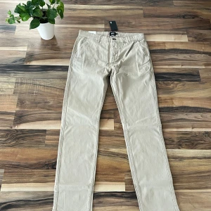Beige chinos från 157, modell Grim - Snygga beige chinos från 157 i modellen Grim. Byxorna har klassisk rak passform med normal midja och är tillverkade i mjuk bomull. Perfekta för en clean och stilren look. Fickor fram och bak, bälteshällor och knappstängning.