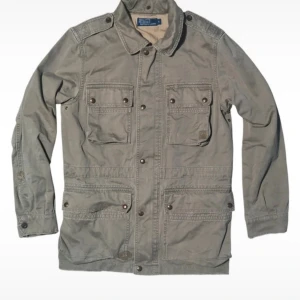 Ralph lauren military jacket - Snygg grå parkajacka från Polo Ralph Lauren i 100% bomull. Jackan har fyra stora fickor med knappar framtill, axelklaffar och en dragkedja på ryggen. Klassisk krage och långärmad design, perfekt för dig som gillar utility-vibe. Size small, men skulle säga att den passar size large.