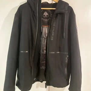 Svart vindjacka Moose Knuckles - Svart  Moose Knuckles jacka (grayton model)  med huva, vattentäta dragkedjor och diskret logga på bröstet. Jackan har slät yta, innerfoder med röda detaljer och två stora fickor framtill. 