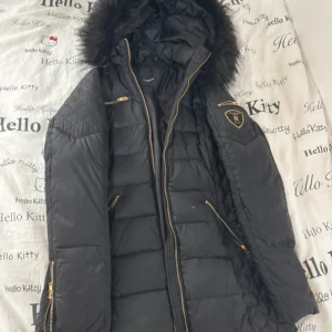 Svart dunjacka Rockandblue med päls - Svart dunjacka från Rockandblue med snygga guldfärgade dragkedjor och quiltade detaljer. Jackan har en stor huva med päls, flera fickor med dragkedja och Rockandblue-logga på ärmen. Perfekt för kalla dagar och riktigt stilren look.