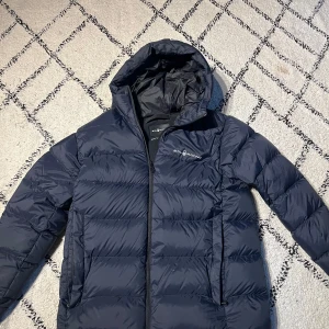 Mörkblå dunjacka från Sail Racing - Snygg mörkblå dunjacka från Sail Racing med huva och dragkedja. Jackan är quiltad och har två sidofickor med dragkedja. Perfekt för kalla dagar och har en stilren look med logga på bröstet. Fodrad insida och klassisk passform.