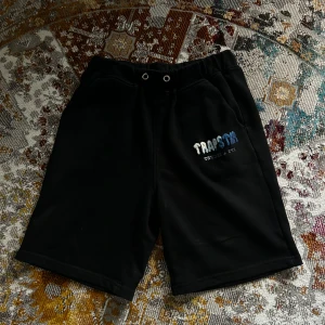 Svarta Trapstar shorts med brodyr - Svarta shorts från Trapstar med broderad logga i vitt och blått på ena benet. Modellen har elastisk midja med snörning och två sidofickor. Perfekta för chill dagar eller streetwear-stil.