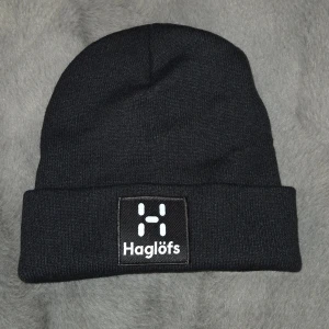 Svart Haglöfs mössa med logga - Svart mössa från Haglöfs med uppvikt kant och tydlig vit logotyp framtill. Klassisk och enkel design som passar perfekt för kalla dagar. Tillverkad i mjukt stickat material som håller dig varm och bekväm.