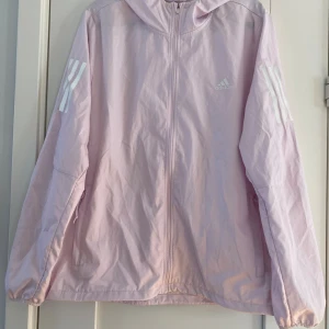 Ljusrosa vindjacka från Adidas - Ljusrosa vindjacka från Adidas med huva och hel dragkedja framtill. Jackan har vita Adidas-ränder på ärmarna och logga på bröstet. Tillverkad i lätt polyester, perfekt för blåsiga dagar. Fickor med dragkedja och elastiska muddar vid ärmslut och nederkant. Mått Över bröstet: ca 58 cm Längd från axeln och ner: ca 65 cm  100% polyester   Kommer från ett djur och rökfritt hem. 