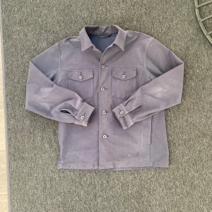 Svart Overshirt från uniqlo - Snygg svart overshirt med klassisk krage och knäppning framtill. Två bröstfickor med lock och knappar ger en clean look. Tillverkad i mjuk bomull, perfekt att slänga över en hoodie eller t-shirt. Långärmad och stilren design. 