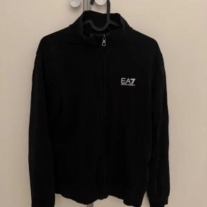 Svart zip-hoodie EA7 Emporio Armani - Svart jacka från EA7 Emporio Armani med hel dragkedja framtill och vit logga på bröstet. Hög krage och diskreta vita ränder på ryggen. Tillverkad i mjukt material, perfekt för en clean och sportig look.
