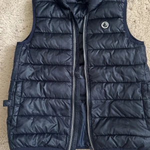 Mörkblå quiltad dunväst Petit Bateau - Snygg mörkblå dunväst från Petit Bateau med quiltad design och dragkedja framtill. Västen har ett stilrent Petit Bateau-logo på bröstet och är tillverkad i polyester. Perfekt för lager-på-lager under höst och vinter.