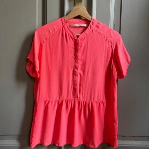 Korallrosa blus från Custommade - Korallrosa blus från Custommade med kort ärm och lätt struktur i tyget. Blusen har en avslappnad passform, liten ståkrage och dold knappslå framtill. Mindre tecken på användning på ena ärmen. 