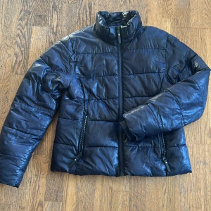 Svart pufferjacka från H&M - Svart pufferjacka från H&M i storlek 152. Jackan har hög krage, två dragkedjeförsedda fickor och quiltad design. Materialet är glansigt och känns lätt men ändå varmt, perfekt för kyliga dagar. Stängs med dragkedja framtill.