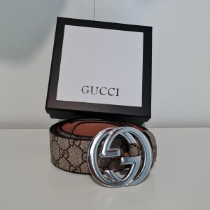 i GG Supreme bälte med silverdetalj - Säljer ett GG Supreme bälte i beige och brunt med klassiskt monogrammönster och stor silverfärgad GG-spänne. Bältet är gjort i äkta skinn och har exklusiv känsla, perfekt för att lyfta vilken outfit som helst.