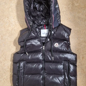 Svart dunväst från Moncler med huva - Snygg svart dunväst från Moncler med glansig finish, stor huva med dragsko och dragkedja framtill. Västen har två fickor med dragkedja och klassisk Moncler-logga på bröstet. Perfekt för lager-på-lager och streetstyle.