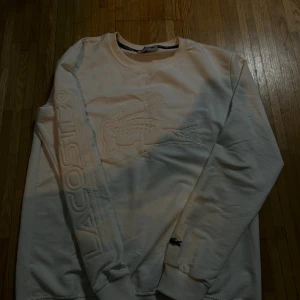 Vit sweatshirt från Lacoste med broderi - Säljer en fräsch vit sweatshirt från Lacoste med stort broderat krokodilmärke på bröstet och broderad Lacoste-text längs ena ärmen. Tröjan har rund hals och långa ärmar med ribbade muddar. Perfekt för dig som gillar stilrena och sportiga plagg.
