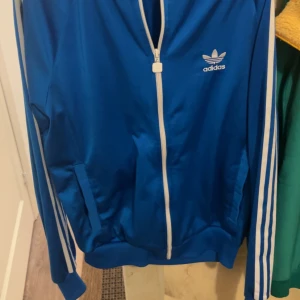 Blå Adidas vindjacka med vita ränder - Klassisk blå Adidas vindjacka med vita dragkedjor och de ikoniska tre ränderna längs ärmarna. Jackan har ståkrage, två fickor och Adidas logga broderad på bröstet. Perfekt för en sportig och stilren look. Använt 3 gånger fortfarande i jätte bra skick 8/10 .