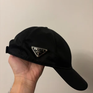 Svart keps från Prada med metallogo - Snygg svart keps från Prada med den ikoniska trekantiga metalloggan på sidan. Kepsen har justerbar rem bak och är tillverkad i ett glansigt syntetmaterial som ger en lyxig känsla. Perfekt accessoar för att lyfta din streetwear-look.