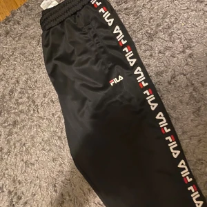 Svarta träningsbyxor från FILA - Svarta träningsbyxor från FILA med elastisk midja och logotyp längs sidorna. Byxorna har en sportig look med vita och röda detaljer i loggan och är tillverkade i ett glansigt syntetmaterial. Perfekta för en aktiv stil.