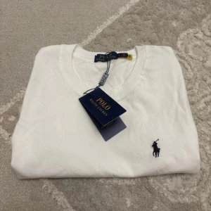Vit t-shirt från Polo Ralph Lauren - Vit t-shirt från Polo Ralph Lauren med klassisk rund hals och svart broderad logga på bröstet. Tillverkad i mjuk bomull och har en normal passform. Perfekt basic-plagg med stilren look och diskreta detaljer.