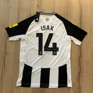 Newcastle United Isak 14 Adidas tröja - Säljer en officiell Newcastle United matchtröja från Adidas med Isak 14-tryck på ryggen. Tröjan är svartvit med klassiska ränder, klubbmärke på bröstet och sponsorlogga framtill. Tillverkad i lätt och ventilerande polyester med AEROREADY-teknologi.