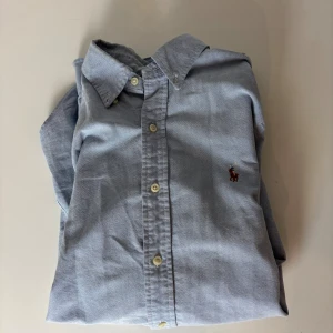 Blå skjorta från Polo Ralph Lauren - Klassisk ljusblå skjorta från Polo Ralph Lauren med broderad logga på bröstet. Skjortan har lång ärm, knappar framtill och button-down krage. Tillverkad i mjuk bomull som känns skön mot huden. Perfekt för en clean och stilren look.