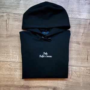 Svart hoodie från Polo Ralph Lauren - Svart hoodie från Polo Ralph Lauren med vit broderad logga på bröstet. Klassisk huva med snörning och mjukt material som känns skönt mot huden. Perfekt för en avslappnad och stilren look.