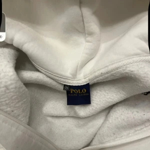 Vit hoodie från Polo Ralph Lauren - Vit hoodie från Polo Ralph Lauren med dragkedja, snörning i huvan och klassisk blå logga broderad på bröstet. Mjuk insida och känguruficka framtill. Perfekt för chill dagar och snygg streetstyle.