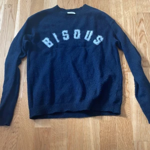 Mörkblå stickad tröja från Kappahl - Mysig mörkblå stickad tröja från Kappahl med texten 'BISOUS' i fluffiga vita bokstäver över bröstet. Tröjan har rund halsringning och långa ärmar, perfekt för kyliga dagar.