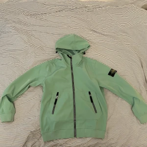 Stone Island Vindjacka - Storlek L - Mintgrön softshelljacka från Stone Island med huva och svarta dragkedjor. Jackan har klassisk patch på ärmen och är tillverkad i stretchig polyester med vindtät och vattenavvisande teknologi. Perfekt för dig som vill ha en clean och sportig look.