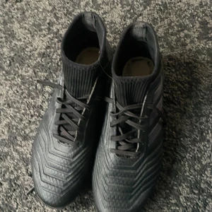 Adidas Predator svarta fotbollsskor - Svarta Adidas Predator fotbollsskor med snörning och ribbad ovandel. Skorna har en högre strumpliknande krage och diskreta detaljer i guld. Tillverkade i syntetmaterial för bästa grepp och kontroll på planen. Obs ingen sula