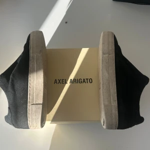 Svarta sneakers från Axel Arigato - Svarta sneakers från Axel Arigato med ovandel i mocka och vit platt sula. Klassisk och stilren design utan snörning, perfekt för dig som gillar minimalistiska vibes. Snygga detaljer och diskret logga på sulan. Boxen medföljer.
