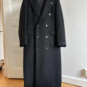 Eytys Genesis kappa i svart - Ullkappa ifrån Eytys modell Genesis i svart. Köpt från Farfetch förra vintern men har bara använt den 2-3 gånger då den inte passar mig.  Den ser ut att vara i nyskick. Relaxed fit, bröstficka samt två djupare fickor. Kappan är 75% ull och 25% polyamid. En avtagbar Eytys pin sitter på den. Den säljs fortfarande på Eytys hemsida för 6900 kr. Storlek women’s EU36 / men’s EU46.