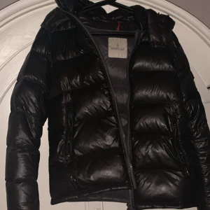 Svart Moncler pufferjacka med huva - Säljer en svart pufferjacka från Moncler med glansig finish och huva. Jackan har dragkedja framtill, två fickor med dragkedja och en extra ficka med Moncler-logga på ärmen. Perfekt för kalla dagar och riktigt snygg streetstil. Sliten med defekter som små små hål vid armen som inte är synliga om man inte kollae noggrant.