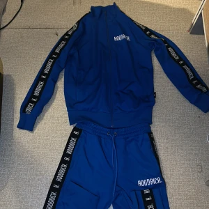 Blå tracksuit från Hoodrich - Säljer en blå tracksuit från Hoodrich med svarta och vita logoband längs ärmarna. Jackan har hel dragkedja framtill, hög krage och långa ärmar. Materialet är mjukt och sportigt, perfekt för en streetwear-look.