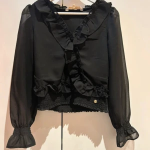 Svart blus med volanger och smock - Supersnygg svart blus med transparenta långärmar och bred smock vid midjan och ärmslut. Volangdetaljer längs halsringningen och framtill. Liten guldfärgad knappdetalj vid midjan. Från Pernilla Wahlgrens kollektion. Använd ett fåtal gånger.       Storlek 34